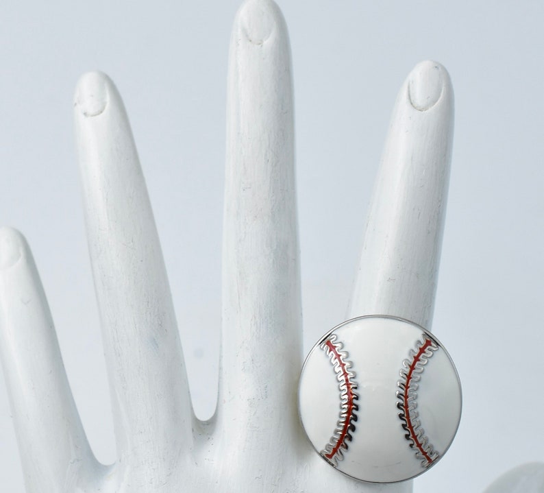 May include: A baseball-themed ring featuring a white enamel design with red stitching and silver accents. The ring is round and sits on a white hand, showcasing the details of the baseball design. The ring is a sports-themed accessory.