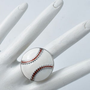 May include: A baseball-themed ring featuring a white enamel design with red stitching and a silver-tone border. The ring is round and resembles a baseball. The ring is shown on a white hand.