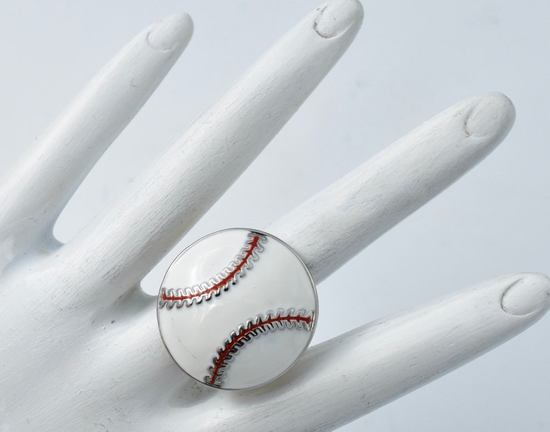 May include: A baseball-themed ring featuring a white enamel design with red stitching and a silver-tone border. The ring is round and resembles a baseball. The ring is shown on a white hand.
