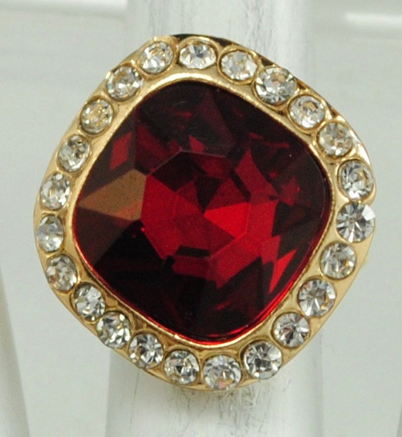 Peut inclure: Une bague dor&eacute;e orn&eacute;e d'une grosse pierre rouge fonc&eacute; taill&eacute;e en forme carr&eacute;e. La pierre est entour&eacute;e de petits cristaux ronds et transparents. La bague est pos&eacute;e sur un fond neutre, avec un design classique.