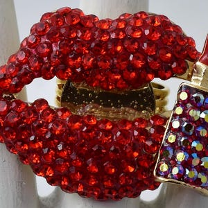 May include: A gold-toned ring featuring a large, red rhinestone-covered lip design. A red lipstick charm with iridescent rhinestones is attached. The ring is a bold, statement piece.