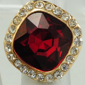 Peut inclure: Bague dor&eacute;e orn&eacute;e d'une grosse pierre rouge fonc&eacute; taill&eacute;e en forme carr&eacute;e. La pierre est entour&eacute;e de petits cristaux ronds et transparents. La monture de la bague est dor&eacute;e.