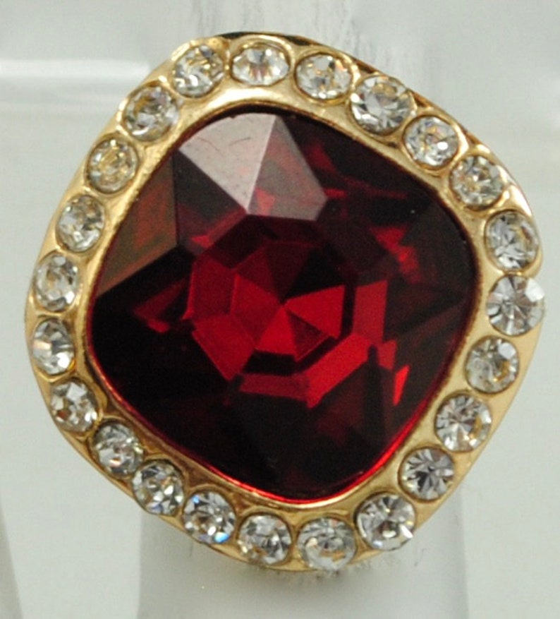 Peut inclure: Bague dor&eacute;e orn&eacute;e d'une grosse pierre rouge fonc&eacute; taill&eacute;e en forme carr&eacute;e. La pierre est entour&eacute;e de petits cristaux ronds et transparents. La monture de la bague est dor&eacute;e.