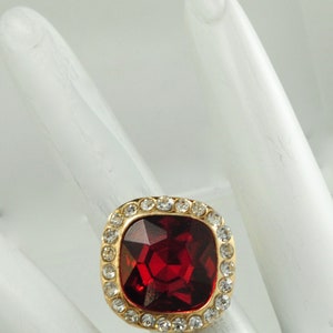 Peut inclure: Bague dor&eacute;e orn&eacute;e d'une grosse pierre rouge carr&eacute;e et facett&eacute;e. La pierre est entour&eacute;e d'un halo de petits cristaux clairs. La bague est pos&eacute;e sur une surface blanche. Bijou de style.
