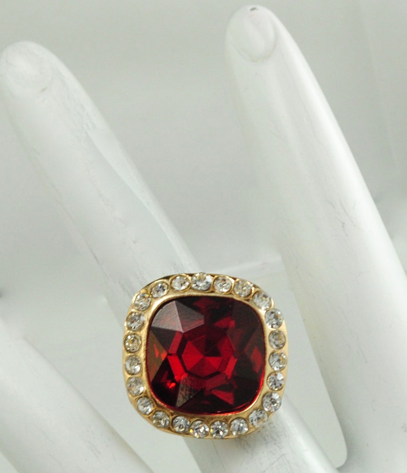 Peut inclure: Bague dor&eacute;e orn&eacute;e d'une grosse pierre rouge carr&eacute;e et facett&eacute;e. La pierre est entour&eacute;e d'un halo de petits cristaux clairs. La bague est pos&eacute;e sur une surface blanche. Bijou de style.