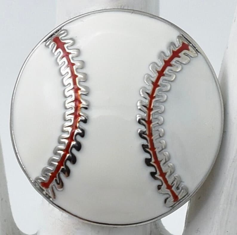 May include: A round baseball-themed ring with a white enamel surface and red stitching detail. The ring has a silver-colored metal border. The baseball design is centered on the ring's face, creating a sporty accessory.