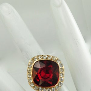 Peut inclure: Bague dor&eacute;e orn&eacute;e d'une grosse pierre rouge carr&eacute;e et facett&eacute;e. La pierre est entour&eacute;e de petits cristaux ronds et transparents. Le bijou est pos&eacute; sur un fond blanc, mettant en valeur son &eacute;clat.