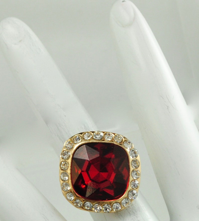 Peut inclure: Bague dor&eacute;e orn&eacute;e d'une grosse pierre rouge carr&eacute;e et facett&eacute;e. La pierre est entour&eacute;e de petits cristaux ronds et transparents. Le bijou est pos&eacute; sur un fond blanc, mettant en valeur son &eacute;clat.