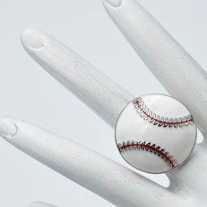 May include: A baseball-themed ring featuring a white enamel design with red stitching details. The ring has a silver-colored metal band and is set against a white background.