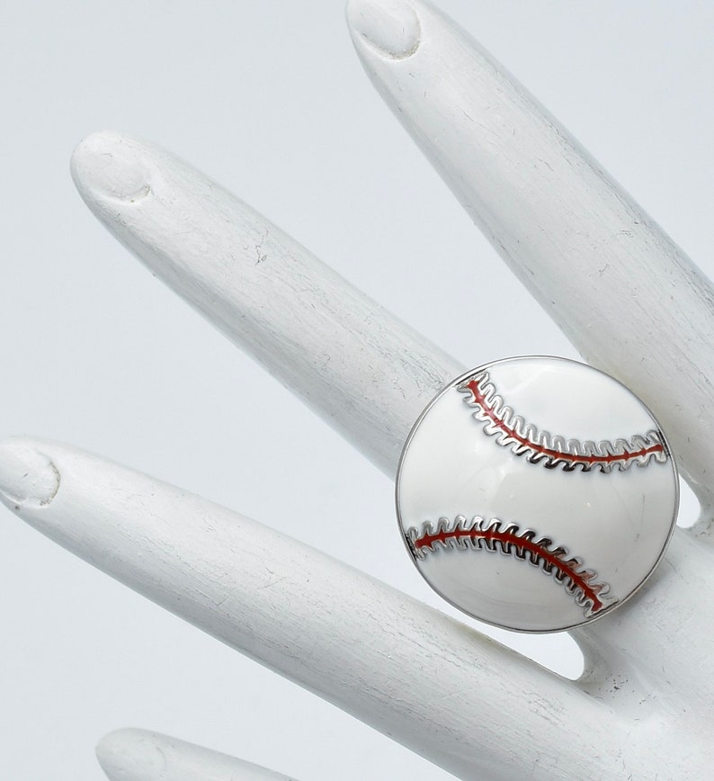 May include: A baseball-themed ring featuring a white enamel design with red stitching details. The ring has a silver-colored metal band and is set against a white background.