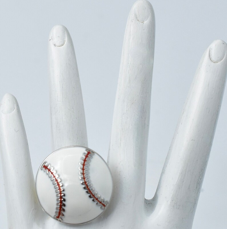 May include: A white baseball ring with red stitching and silver accents. The ring is round and has a glossy finish, resembling a miniature baseball. The ring is a sports-themed accessory.