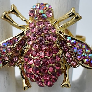 May include: A gold-toned ring featuring a large insect-shaped centerpiece. The body and wings of the insect are encrusted with numerous round, pink faceted crystals that catch the light, creating a sparkling effect across the entire surface.