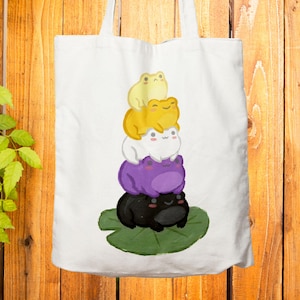 May include: White tote bag with a design of stacked cartoon frogs in yellow, gold, white, purple, and black. The frogs are on a green lily pad. The bag has two white handles and is set against a wooden background.