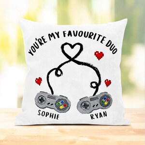 May include: White square cushion with the text "YOU'RE MY FAVOURITE DUO" in black. Two grey game controllers are linked by a black heart-shaped cable. The names "SOPHIE" and "RYAN" are below the controllers, with red pixelated hearts.