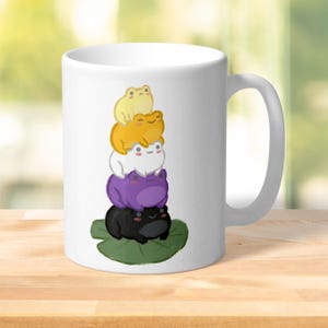 May include: White ceramic mug with a design of cartoon frogs in yellow, orange, white, purple, and black, stacked on a green lily pad. The mug has a white handle.