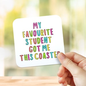 May include: A white square coaster with rounded corners, held by a hand. The coaster features the text "MY FAVOURITE STUDENT GOT ME THIS COASTER" in colourful bubble letters. The background is blurred.