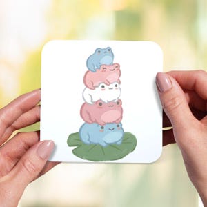 May include: A square white coaster with a design of cartoon frogs in blue, pink, and white. The frogs are stacked, with the bottom frog on a green lily pad. The coaster is held by two hands. The design is simple and colourful.