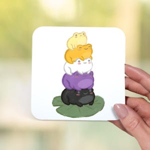 May include: A white square sticker featuring a stack of five cartoon frogs. The frogs are yellow, orange, white, purple, and black, stacked on a green lily pad. The frogs have smiling faces.