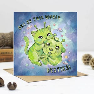 May include: Greeting card with two green alien-like creatures, a cat and a dog, with antennae. The card reads "OUT OF THIS WORLD BESTIES!" against a blue and purple watercolour background with stars and hearts.