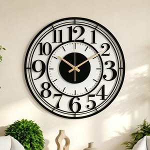 May include: A large circular wall clock featuring a black metal frame with bold black numerals from one to twelve. The clock face has a white background with a central black circle and thin gold-colored hands for the hour, minute, and second.