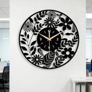 Puede incluir: Un reloj de pared redondo negro con un diseño floral. La esfera del reloj es negra con manecillas y números dorados. El borde exterior presenta flores y hojas recortadas, creando un borde decorativo. El reloj está montado en una pared blanca.