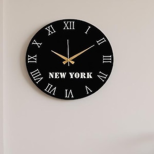 Op de afbeelding: Een zwarte ronde wandklok met witte Romeinse cijfers en gouden wijzers. De klok heeft de tekst "NEW YORK" in het wit. De klok is aan een witte muur bevestigd.
