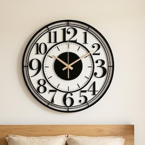 May include: A large round wall clock featuring a black metal frame with oversized black numerals from one to twelve. The clock face has a white background with a central black circle and thin gold-coloured hour, minute, and second hands.