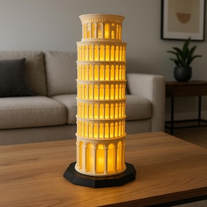 May include: A white ceramic lamp shaped like the Leaning Tower of Pisa with a warm yellow light emanating from the windows. The lamp is sitting on a wooden table.