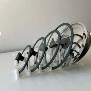 Puede incluir: Un organizador de tapas de ollas de plástico blanco que sostiene varias tapas de ollas de vidrio y metal. Las tapas varían en tamaño y tienen asas negras y bordes grises. El organizador está sobre una superficie blanca.