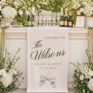 May include: A cream-colored fabric table runner featuring dark green text that reads CHEERS TO The Wilsons HELLEN & STEVE 10.15.2026 with a graphic of two clinking champagne glasses. The runner is draped over a white bar with floral arrangements.