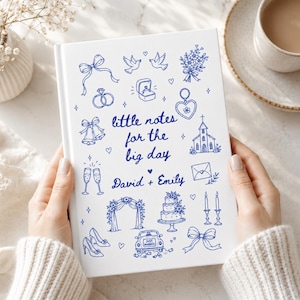 May include: A white hardcover notebook featuring blue line art illustrations of wedding icons including rings, a church, a cake, and a car. The text reads little notes for the big day and David + Emily in a blue script font on the front cover.