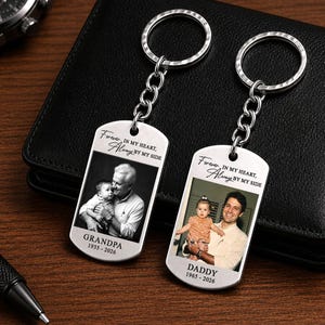 May include: Two silver keychains with photo pendants. Each keychain has a photo of a person holding a baby. One keychain says "Grandpa 1935-2026" and the other says "Daddy 1965-2026". Both keychains have the text "Forever in my heart, Always by my side".