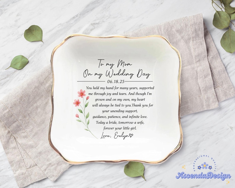 May include: A white square ceramic dish with a gold rim. The dish has a heartfelt message in black script: 'To my Mom On my Wedding Day 06.18.25- You held my hand for many years, supported me through joy and tears... Love, Evelyn.' A floral design with red flowers and green leaves is on the left side. The dish is on a marble surface with a linen napkin.