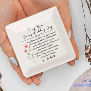 May include: A square, white ceramic trinket dish with gold trim, held in a person's hands. The dish features a floral design and a heartfelt message: "To my Mom On my Wedding Day." The text includes a date and a personal note.