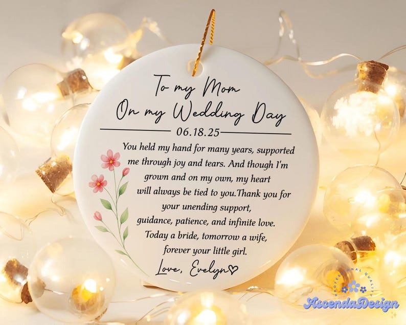 May include: A white ceramic ornament with a gold string for hanging. The ornament has a floral design with pink flowers and green leaves. Text on the ornament reads 'To my Mom On my Wedding Day 06.18.25 You held my hand for many years, supported me through joy and tears. And though I'm grown and on my own, my heart will always be tied to you. Thank you for your unending support, guidance, patience, and infinite love. Today a bride, tomorrow a wife, forever your little girl. Love, Evelyn'.