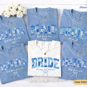 May include: A collection of light blue and white t-shirts with text in a patchwork design. The shirts say "Mother of the Bride", "Maid of Honor", "Bridesmaid", "Bride", and "Mother of the Groom". Each shirt has a gold necklace.