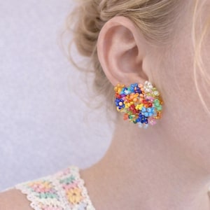May include: A vibrant, beaded earring with a cluster of small beads in shades of red, orange, yellow, blue, and green. The earring is square-shaped and worn on the ear. The background is a pale, neutral tone.