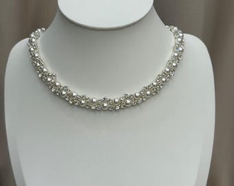 May include: A delicate necklace featuring alternating rows of small, round pearls and sparkling clear crystals. The necklace is displayed on a white mannequin, showcasing its elegant design. The piece is likely intended for formal occasions.