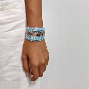 Könnte beinhalten: Ein mehrreihiges Perlenarmband mit Reihen aus hellblauen und metallisch bronzefarbenen Saatperlen. Das Armband besteht aus mehreren horizontalen Bändern, die sich um das Handgelenk legen und ein breites, geschichtetes Erscheinungsbild mit einem durchgehenden Farbmuster erzeugen.