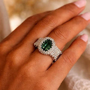 Peut inclure: Une bague en argent ornée d'une pierre précieuse ovale vert émeraude. La bague présente un motif de perles autour de la pierre et sur le corps. La bague est portée sur une main à la peau claire.