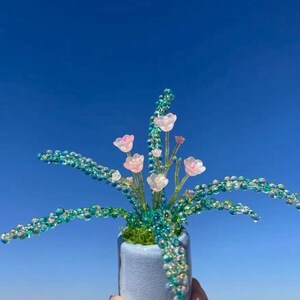 Pode incluir: Um arranjo decorativo com pequenas flores translúcidas em rosa pálido e caules longos e arqueados cobertos por contas cintilantes em tons de azul-petróleo e azul. A planta de contas está inserida num pequeno vaso de cerâmica azul claro.