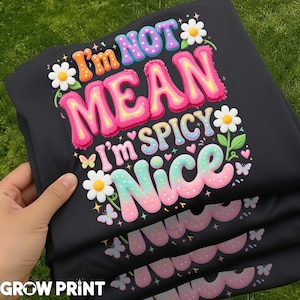 以下が含まれることがあります： 「I'm NOT MEAN I'm SPICY NICE」の文字がカラフルにプリントされた黒いスウェットシャツ。 デイジー、蝶、ハートのイラストがデザインされています。 スウェットシャツは手で持たれており、下部にはGrow Printのロゴが見えます。