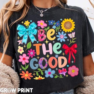 May include: Dark gray t-shirt with the phrase "BE THE GOOD" in colorful, patchwork-style letters. The design includes various flowers, bows, and butterflies in bright colors. The brand name "GROW PRINT" is at the bottom.