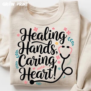 May include: A beige sweatshirt with the text "Healing Hands, Caring Heart!" in black lettering. The text is surrounded by pink and blue flowers and a black and pink stethoscope.