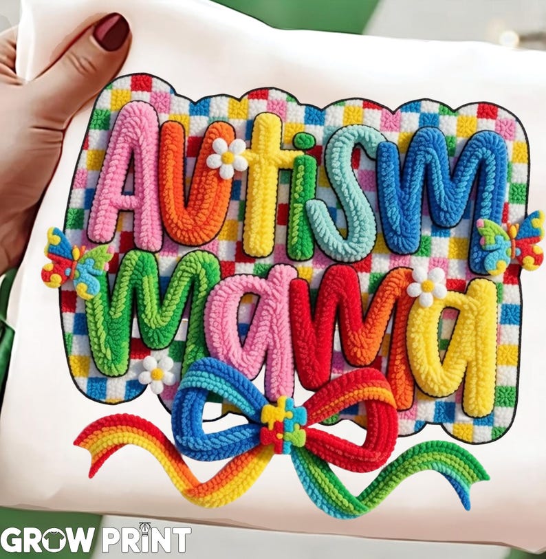 May include: A white t-shirt featuring the words "AUTISM MAMA" in colorful, textured letters. The design includes a rainbow bow, butterflies, and small flower accents. The background is a checkered pattern.
