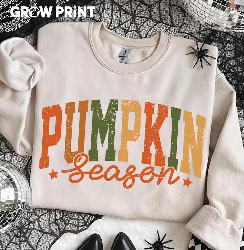 Peut inclure: Sweat-shirt cr&egrave;me avec l'inscription "PUMPKIN Season" en orange, vert et jaune. Le sweat-shirt a un col rond et des manches longues. Le texte "GROW PRINT" est en haut. Des araign&eacute;es d&eacute;coratives et une boule disco sont en arri&egrave;re-plan.