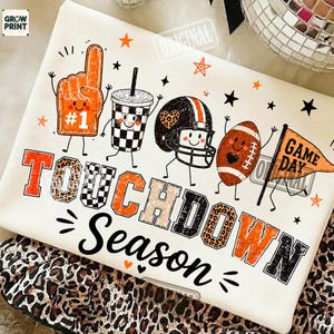 Może przedstawiać: Biały t-shirt z nadrukiem graficznym przedstawiającym piankowy palec, kubek w szachownicę, kask do futbolu amerykańskiego, piłkę oraz proporczyk z napisem GAME DAY. Poniżej znajduje się napis TOUCHDOWN SEASON w kolorze pomarańczowym, czarnym i w cętki.