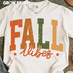 Peut inclure: T-shirt blanc avec l'inscription "FALL vibes" en lettres orange, vertes et jaunes. Le texte a un aspect vieilli et vintage, avec deux petites &eacute;toiles orange. Le t-shirt a un col en V et des manches courtes. La marque "GROW PRINT" est en haut.