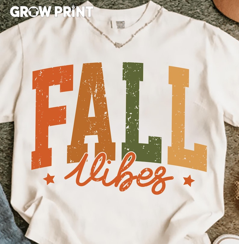 Peut inclure: T-shirt blanc avec l'inscription "FALL vibes" en lettres orange, vertes et jaunes. Le texte a un aspect vieilli et vintage, avec deux petites &eacute;toiles orange. Le t-shirt a un col en V et des manches courtes. La marque "GROW PRINT" est en haut.
