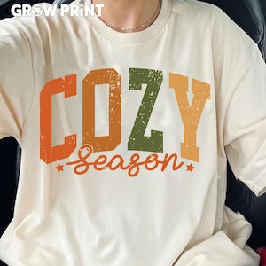 Peut inclure: T-shirt cr&egrave;me avec l'inscription "COZY Season" en lettres d&eacute;lav&eacute;es orange, vert et jaune. Le t-shirt a un col rond et des manches courtes. La marque "GROW PRINT" est en haut.